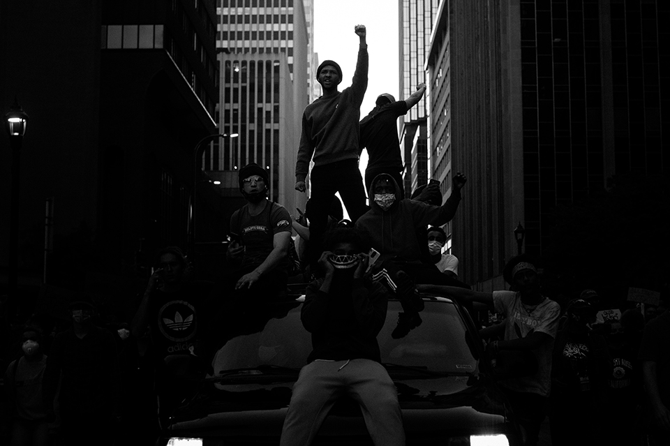 Two protestors stand atop a car, while several others sit on top of the car, with fists in the air in downtown Los Angeles in Brandon Bell's black-and-white photo.