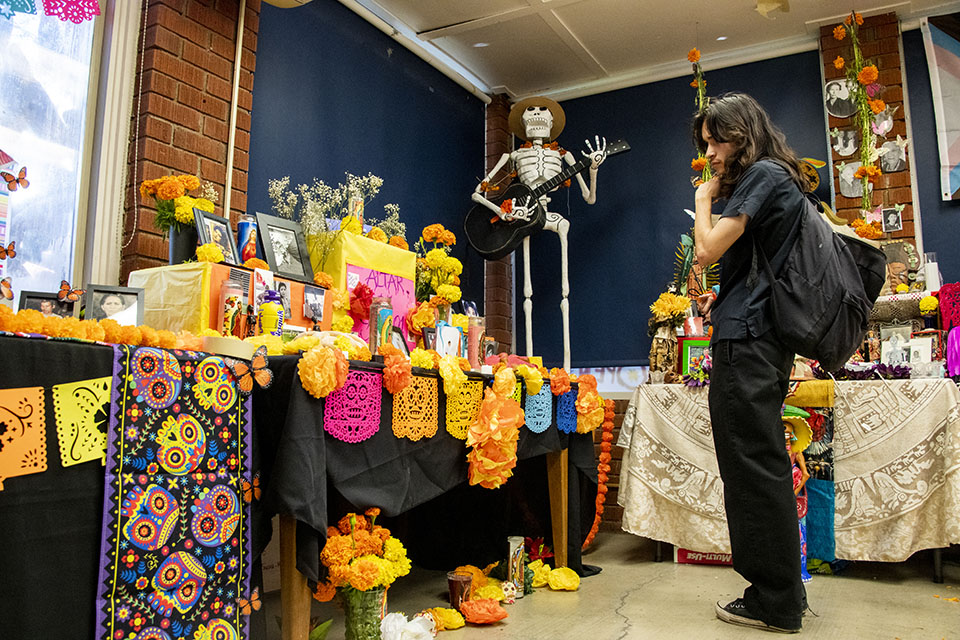 A student dressed all in black stands inside the Chicano House, looking at a large ofrenda (altar) covered in colorful art, papel picado and photos.