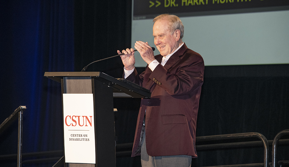 Conference founder Harry Murphy stands on stage alongside podium and gestures to the crowd
