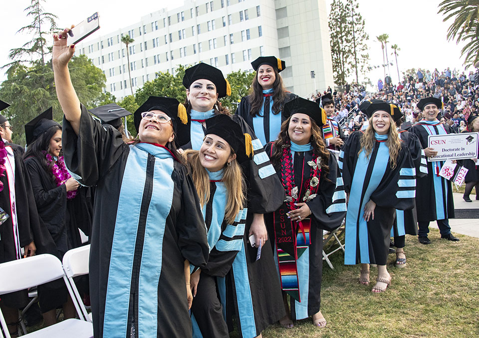 A row of doctoral graduates in black robes and light-blue stripes pose for a group selfie at Commencement 2022.