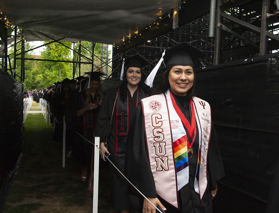 Smiling college graduate is wearing a white stole that says 'CSUN' and arriving to a graduation ceremony.