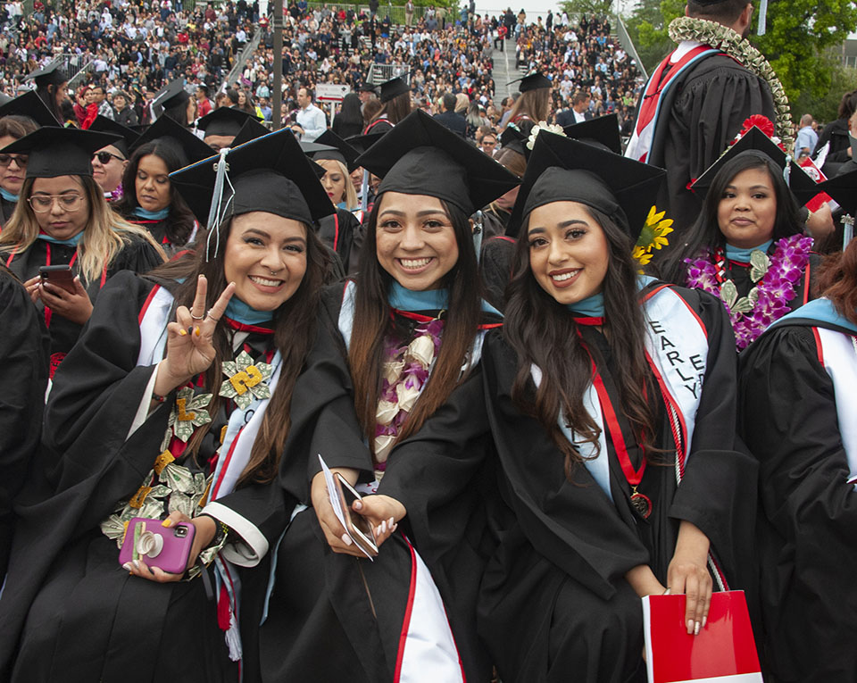group of three smiling college graduates sit and pose together during a graduation ceremony.