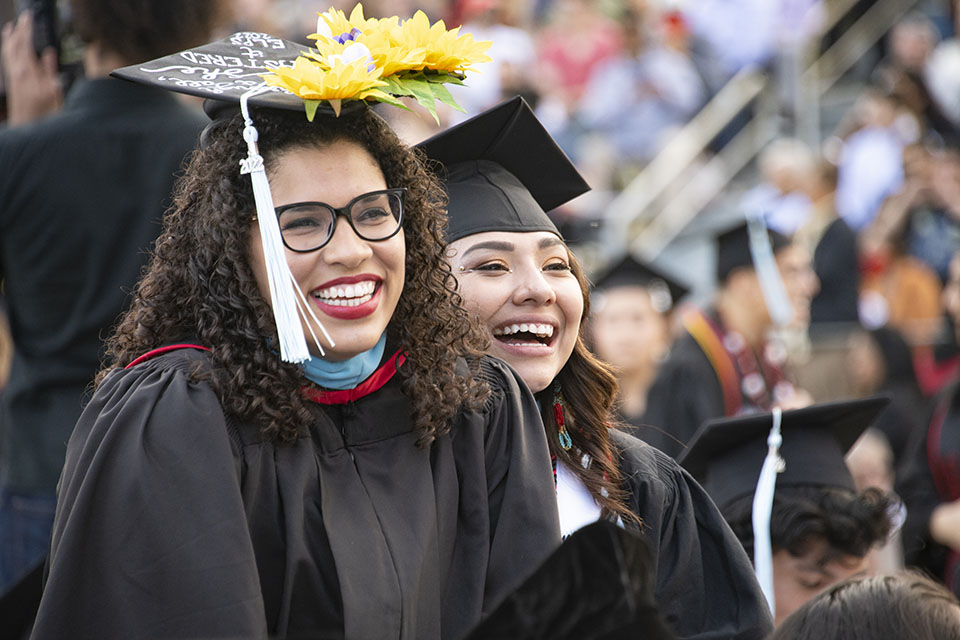 Graduates in brightly decorated caps, gowns and master's degree hoods stand and smile, at their seats at Commencement 2022.