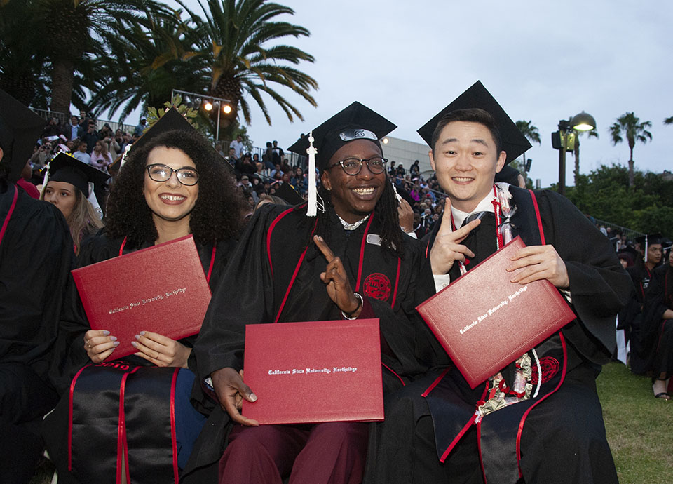 group of three smiling college graduates hold up red diplomas and pose for a camera during a graduation ceremony.