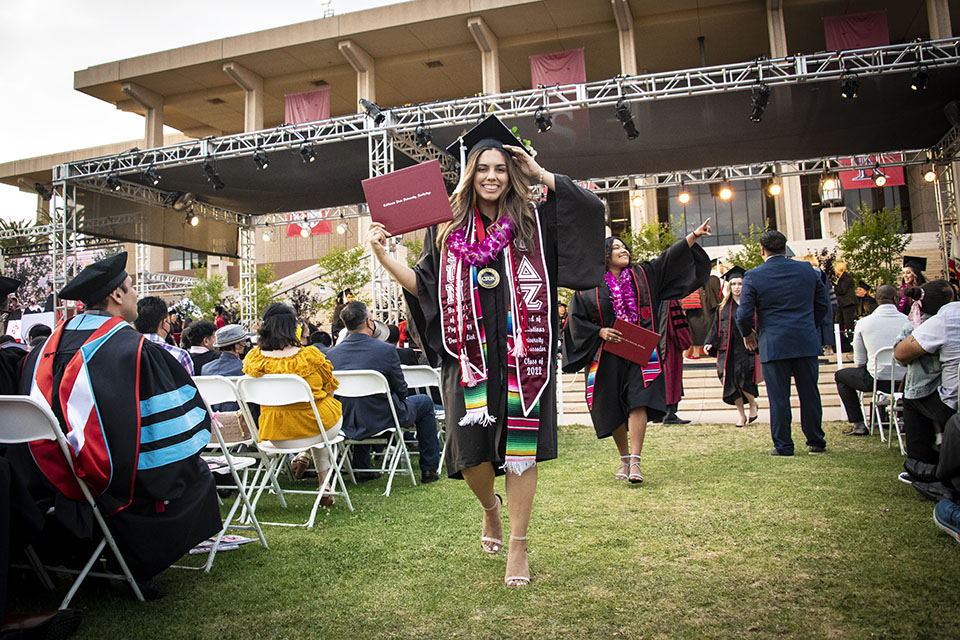 A graduate in cap, gown and sash holds up her red diploma cover as she walks off the stage at Commencement 2022, in front of the University Library.