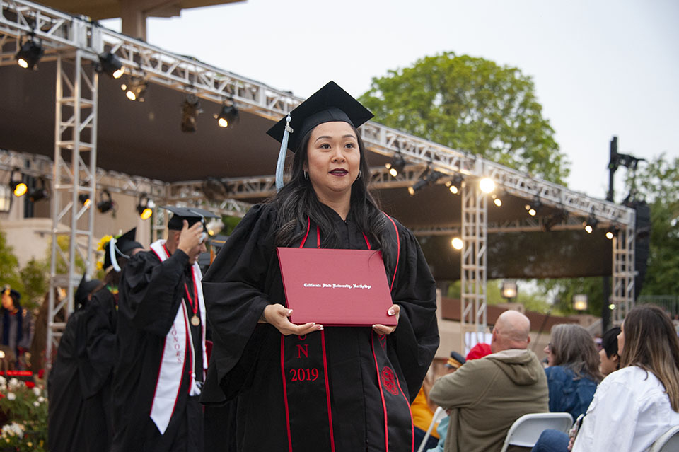 smiling college graduate holds a red diploma and poses for a camera at a graduation ceremony.