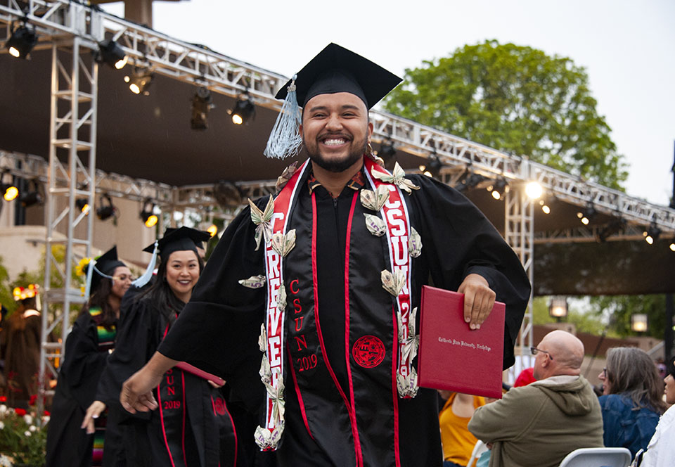 smiling college graduate is wearing a red stole with money pinned onto it and is holding a red diploma at a graduation ceremony.