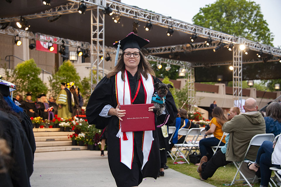 smiling college graduate is holding a red diploma and a small dog at a graduation ceremony.