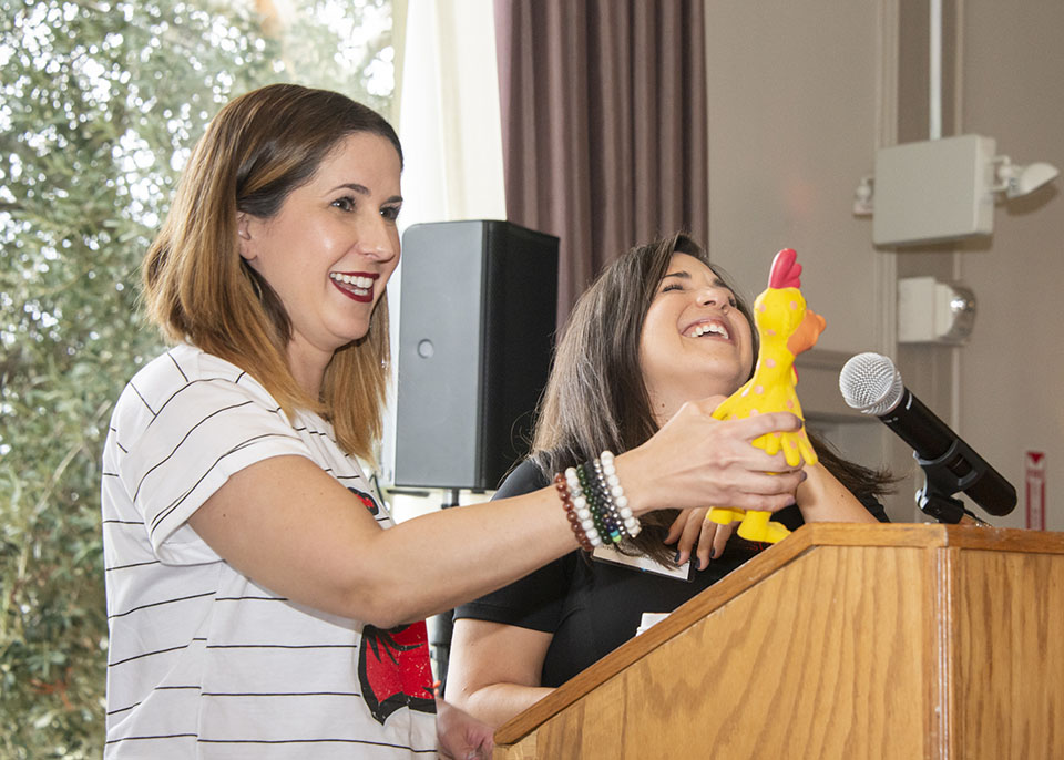 One female professor holds a yellow, rubber chicken squeak toy, while the woman standing next to her laughs. Both are standing at a podium.