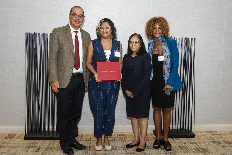 Four people stand together and pose for the camera. Pavithra Prasad stands holding her award in a red folder.