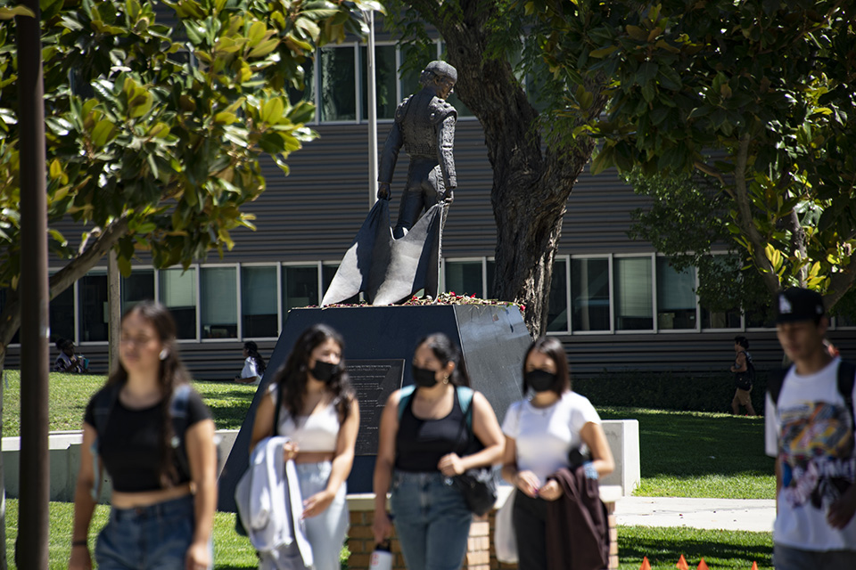 Matador statue in the background with red roses piled on the base of the statue. Students walk on sidewalk in foreground.