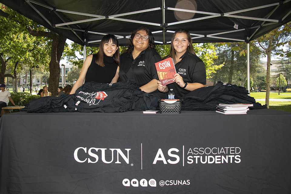 Students wearing black polo shirts stand behind a table with an Associated Students banner, one is holding a planner.