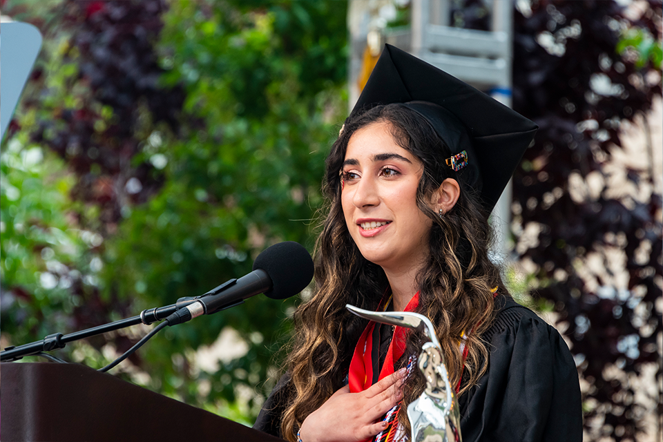 Eden Shashoua in cap and gown stands at the podium on the commencement stage