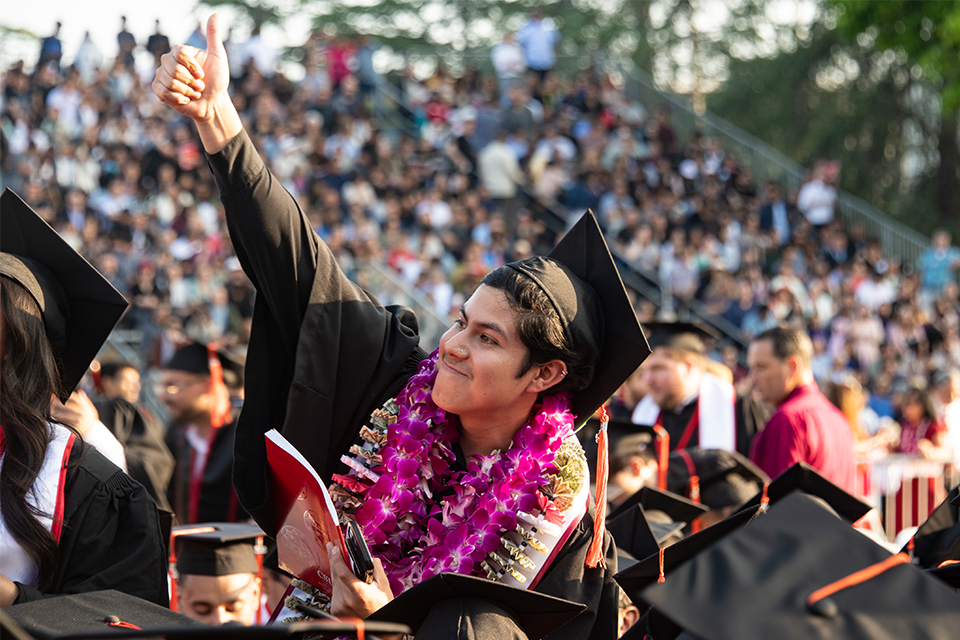 A graduate in cap, and gown and colorful lei emerges up from the seated crowd to give a triumphant wave