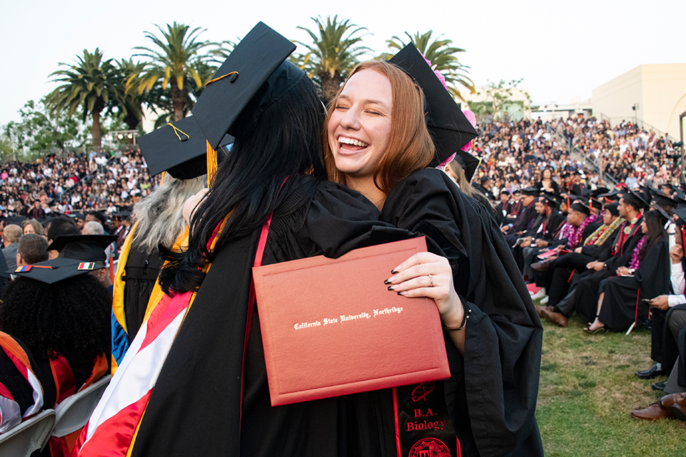 Two women hug, woman facing camera holds out diploma cover