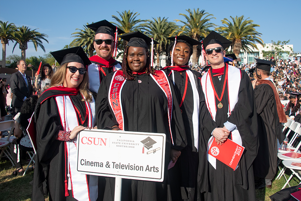 A group of five graduates in cap and gown pose in front of a sign that says Cinema and Television Arts.