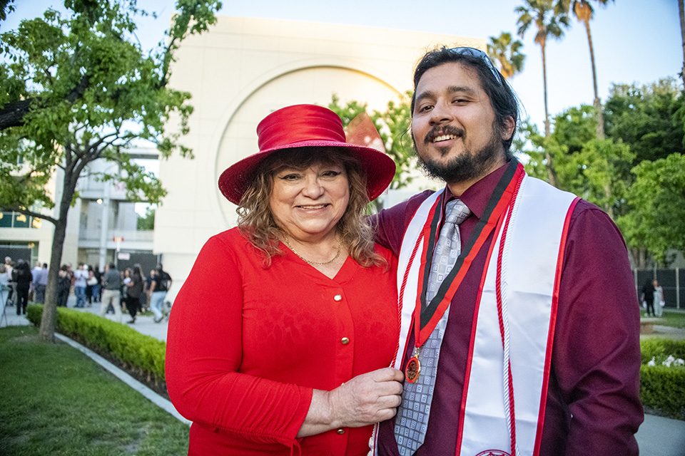 A beaming Mom, dressed in a red sweater and red hat, stands next to her son, who is wearing a white graduation sash and Honors medal.