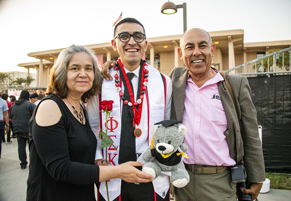A mother and father stand on either side of their son, who is holding a graduation teddy bear and a red rose, in front of the University Library.