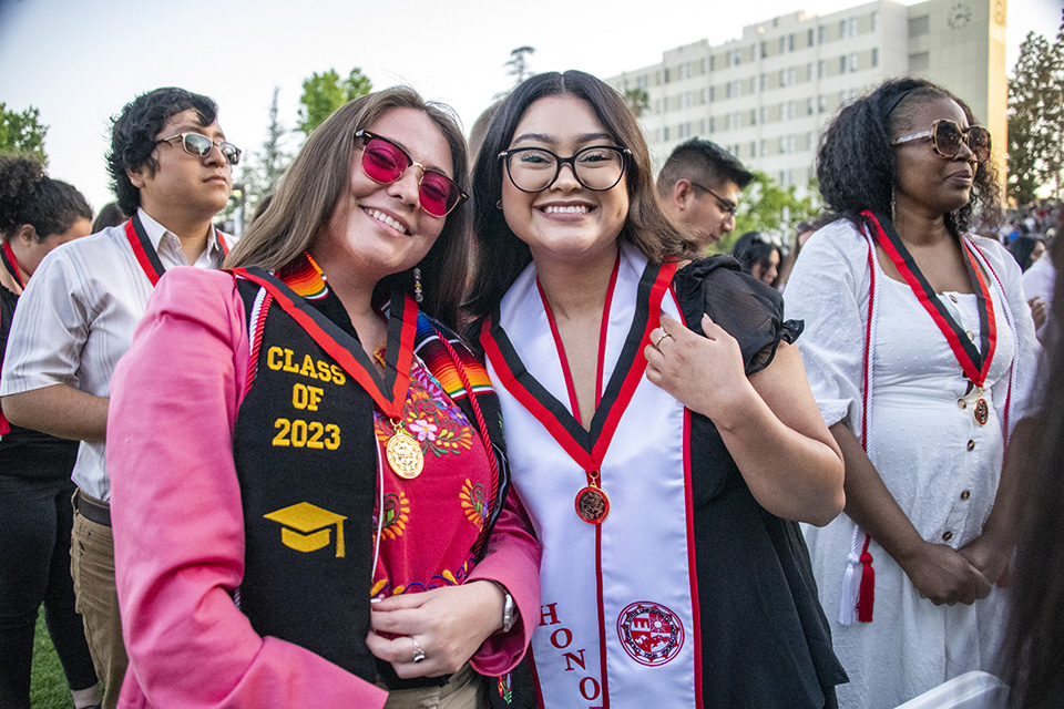 Two graduates, wearing honors medals, hug and smile. Sierra Tower is in the background.