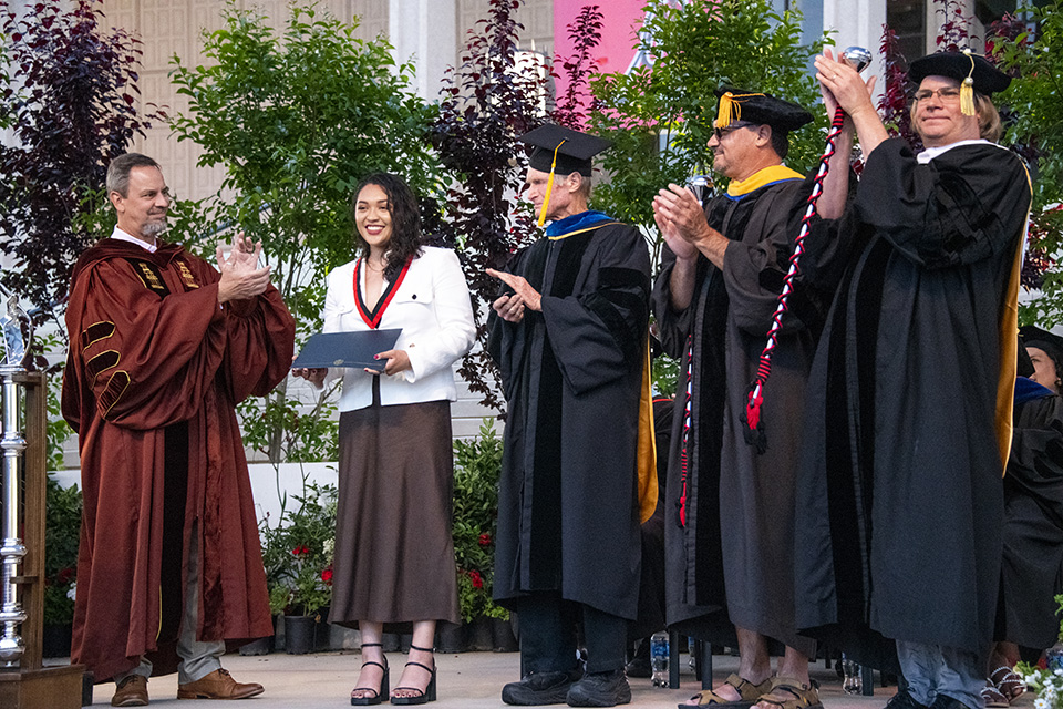 Wolfson Scholar Award winner Natalie Castillo holds her certificate, while deans and faculty members stand next to her on stage and applaud.