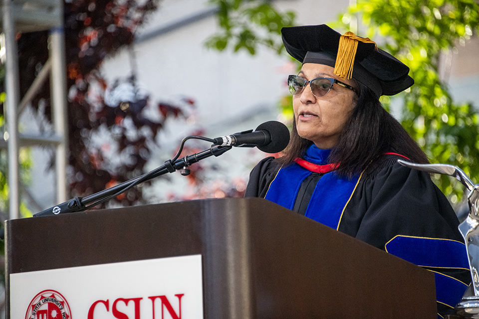 Provost Meera Komarraju, in cap and gown, stands behind a podium and speaks into the microphone.