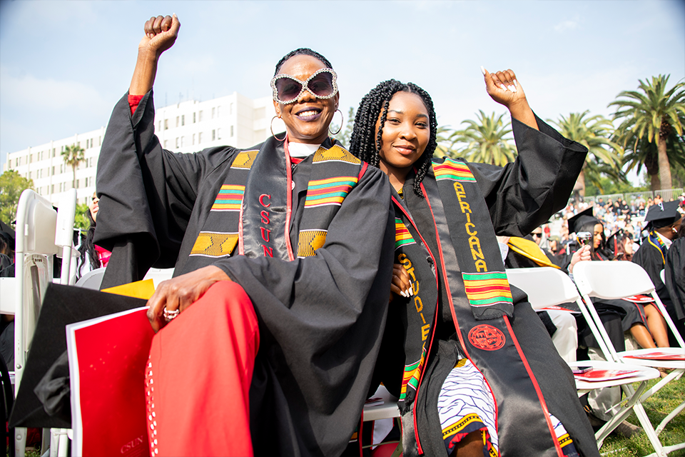 Two women in graduation robes and brightly colored sashes each hold up a fist in triumph and celebration