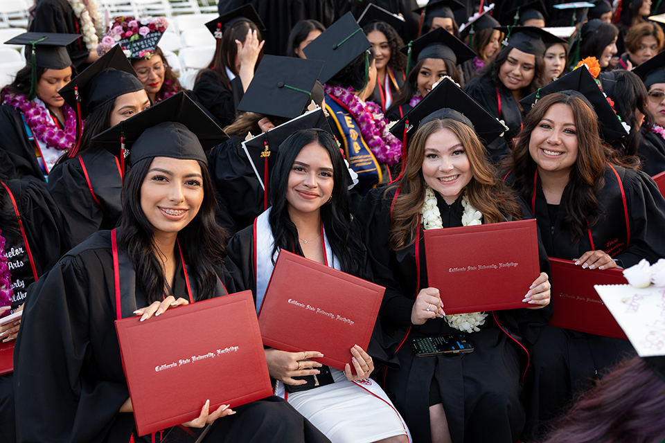 Four women in caps and gowns show their red diploma covers