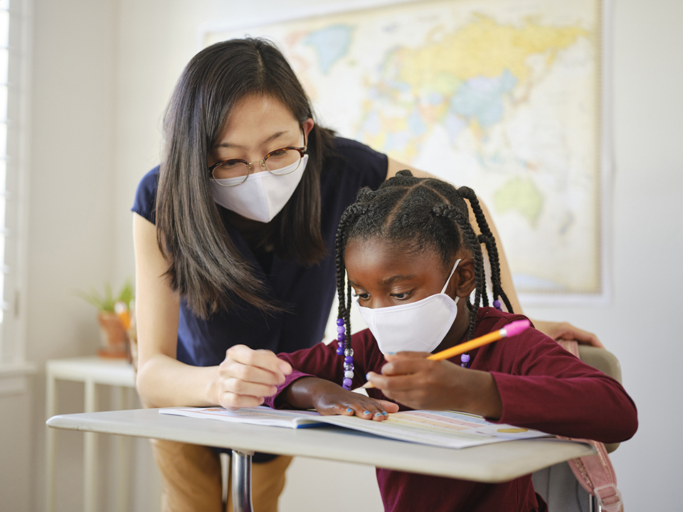 An elementary aged school student and teacher in a classroom wearing a mask for protection against infectious disease.