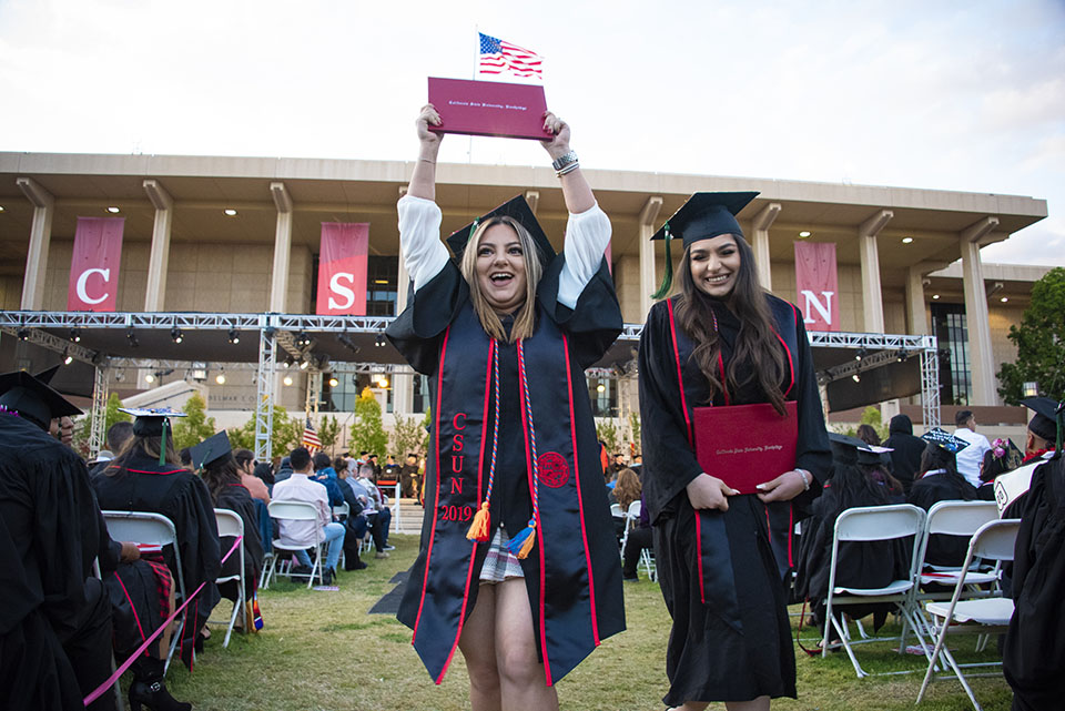 A female graduate hoists her diploma in the air while a fellow graduate hugs hers close.
