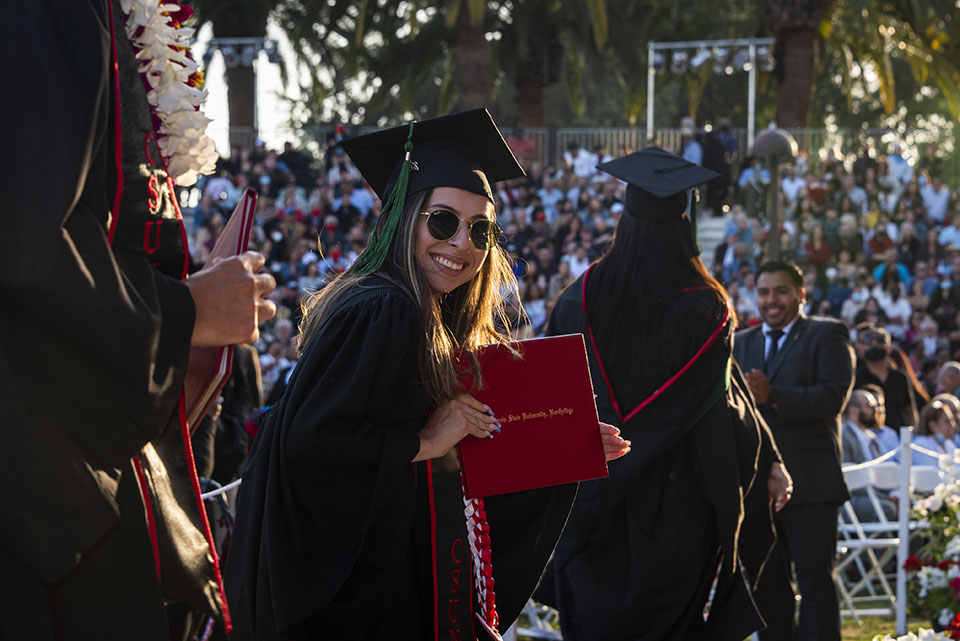A brand-new graduate in cap and gown smiles and holds up her red diploma cover, after walking off the stage at Commencement 2022.