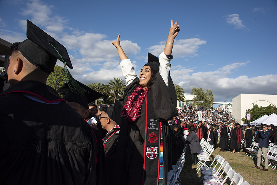 A female graduate raises her arms triumphantly on a sunny day at a CSUN ceremony.