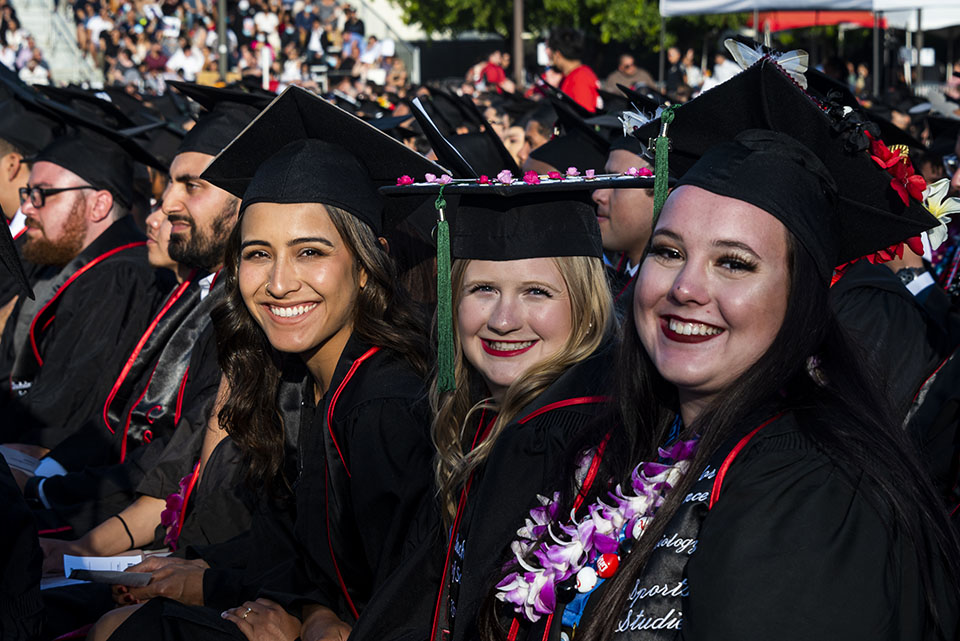 A row of graduates in caps, gowns and flower leis smile and celebrate at Commencement 2022.
