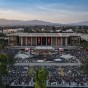 A drone camera aerial view of the University Library and Commencement crowds in bleachers and sections of chairs, with the CSUN campus and north San Fernando Valley beyond.