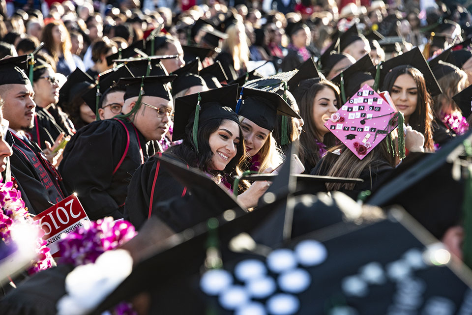 Two females in a sea of seated CSUN graduates lean together for a selfie.