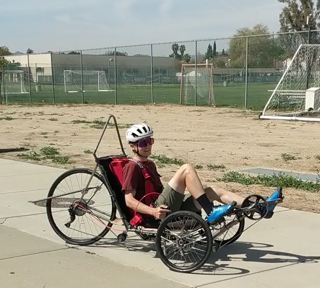 A student sporting sunglasses and a white helmet rides a three-wheeled bike while sitting down.