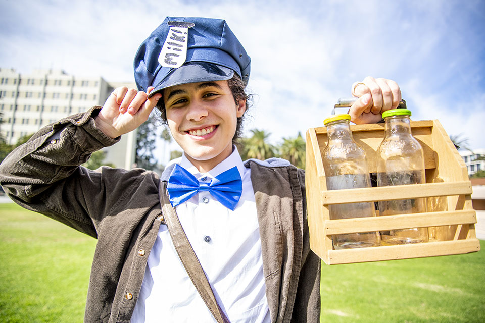 Student wearing blue bow tie and blue cap carrying a wooden crate filled with empty milk bottles