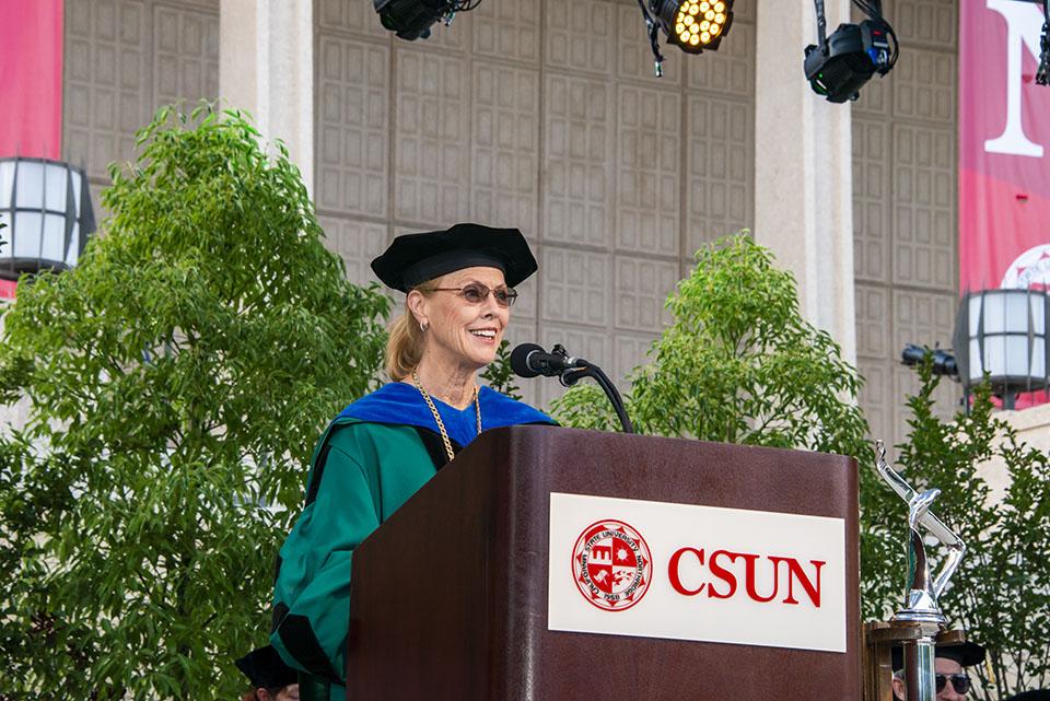 university president dianne f. harrison gives a speech at a wooden podium on stage during a graduation ceremony.