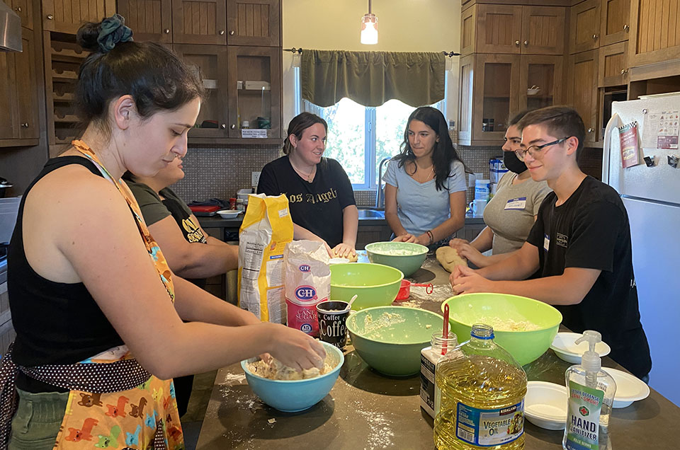 Six CSUN students and Hillel staff members stand around a kitchen island, mixing challah dough with their hands and rolling out dough on the counter.