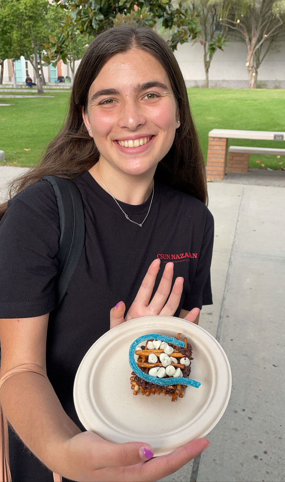 A CSUN student stands on Matador Walk, hoding a paper plate with a candy sukkah built out of a pretzel sticks, marshmallows, frosting and licorice.