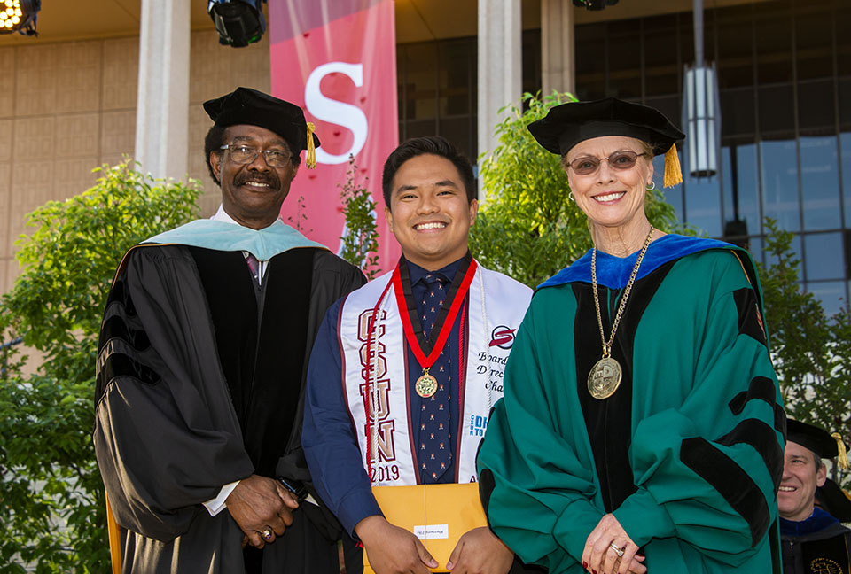 dean of student and vice president for student affairs william watkins and university president dianne f. harrison smile and congratulate smiling outstanding graduating senior award winner bhernard tila who is wearing a medal and holding a yellow envelope on a stage during a graduation ceremony.
