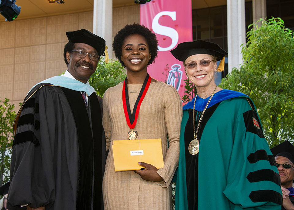 dean of student and vice president for student affairs william watkins and university president dianne f. harrison smile and congratulate smiling outstanding graduating senior award winner and outgoing associated students president beverly ntagu who is wearing a medal and holding a yellow envelope on a stage during a graduation ceremony.