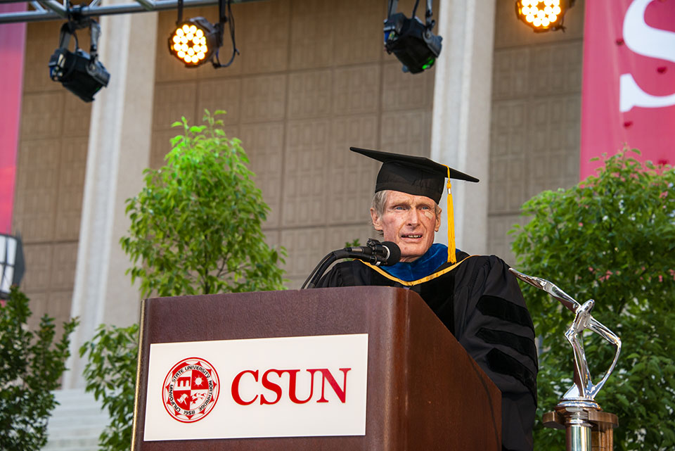 dean of college of sciences and mathematics jerry stinner gives a speech at a wooden podium that says 'CSUN' on stage at a graduation ceremony.