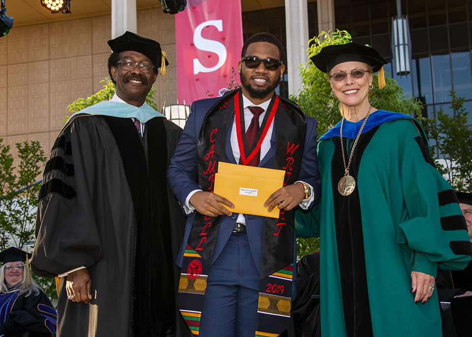 dean of student and vice president for student affairs william watkins and university president dianne f. harrison smile and congratulate smiling outstanding graduating senior award winner khari campbell-wright who is wearing a medal and holding a yellow envelope on a stage during a graduation ceremony.