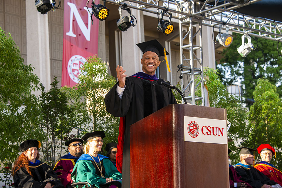alumnus robert d. taylor gives a speech at a wooden podium that says 'sun' on stage at a graduation ceremony.