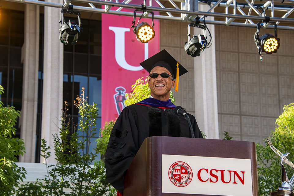 close up shot of alumnus robert d. taylor as he gives a speech at a wooden podium that says 'sun' on stage during a graduation ceremony.