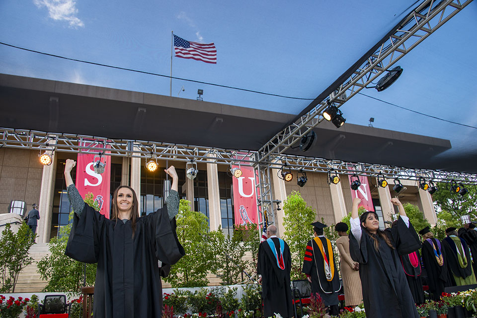 two university students wearing black robes hand sign the national anthems during a graduation ceremony.