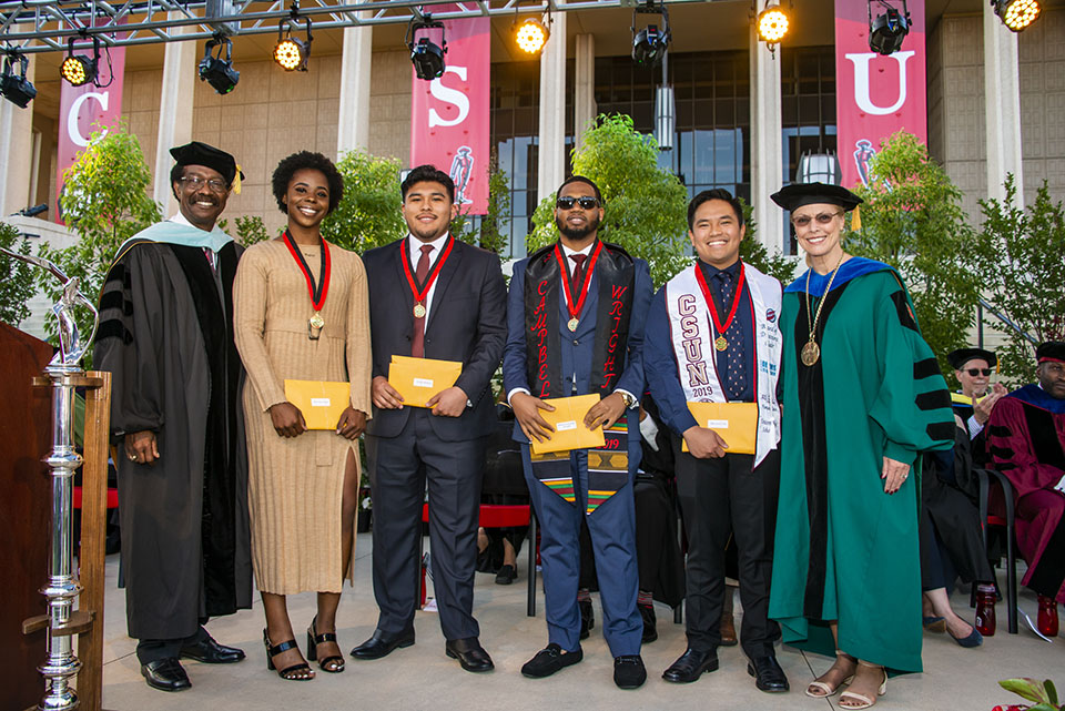 dean of student and vice president for student affairs william watkins and university president dianne f. harrison smile and congratulate outstanding graduating senior award winners who are wearing medals and holding yellow envelopes on a stage during a graduation ceremony.