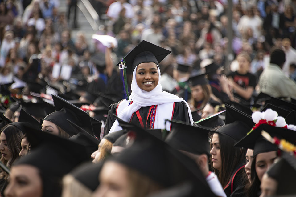 A graduate in black cap and gown and white hijab stands to be recognized at Commencement 2022.