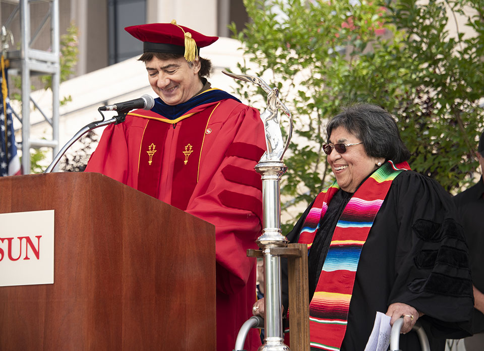 CSUN alumna Irene Tovar stands at a podium, to receive an honorary Doctor of Humane Letters from a member of the CSUN faculty, at Commencement 2022.