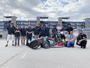 The students and the race car are smiling for the picture, standing in front of a building. 